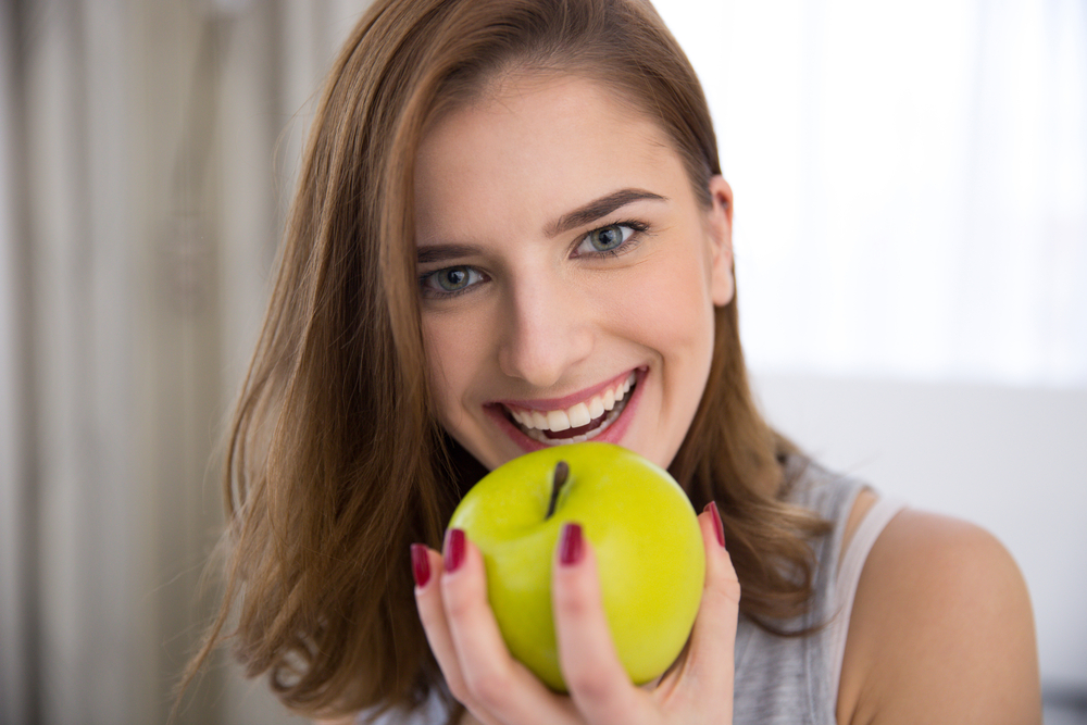 Woman smiling while holding a green apple, demonstrating strong, functional teeth and the benefits of dental bridge restoration - Dental Bridges San Diego
