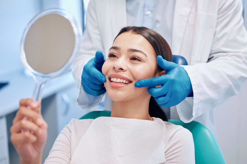Female patient holding a mirror and smiling while dentist adjusts her cheeks, highlighting natural-looking results from dental implant treatment - Dental Implants San Diego