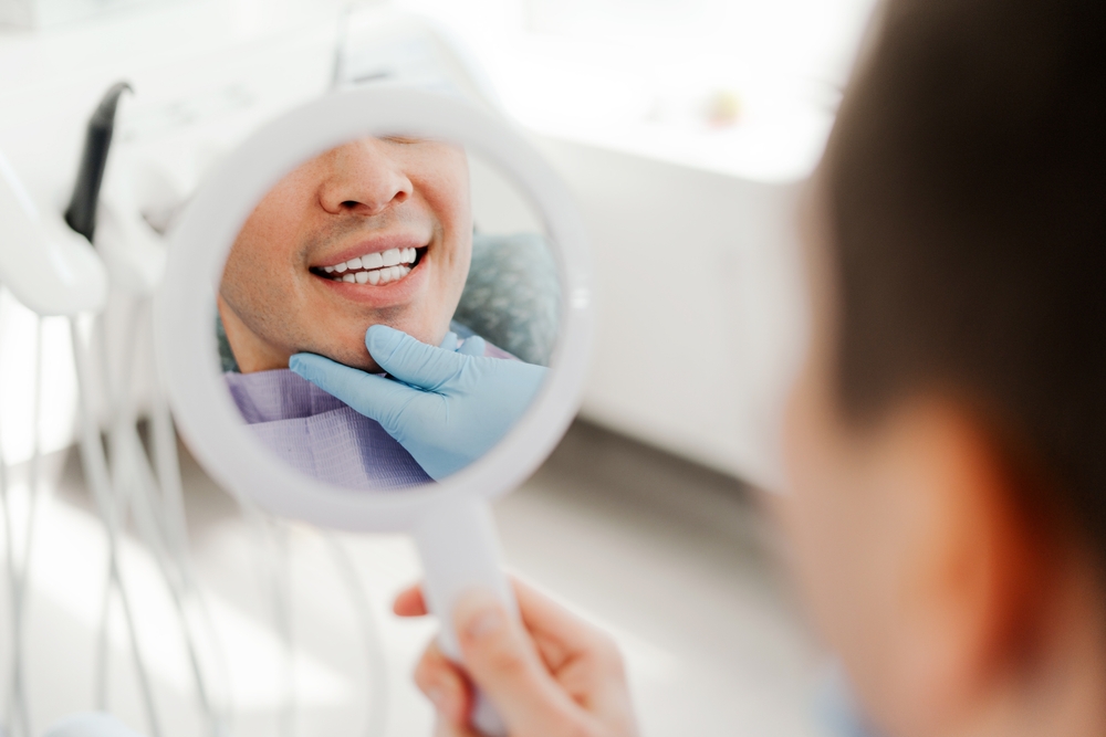 Close-up reflection of a smiling patient in a handheld mirror while dentist examines teeth, showcasing successful dental implant results - Dental Implants San Diego