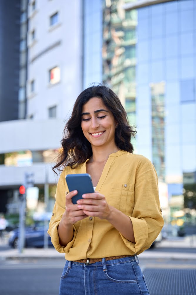 A woman in the city with a yellow blouse smiles as she checks her phone - Truly Yours Family Dental in La Mesa, CA